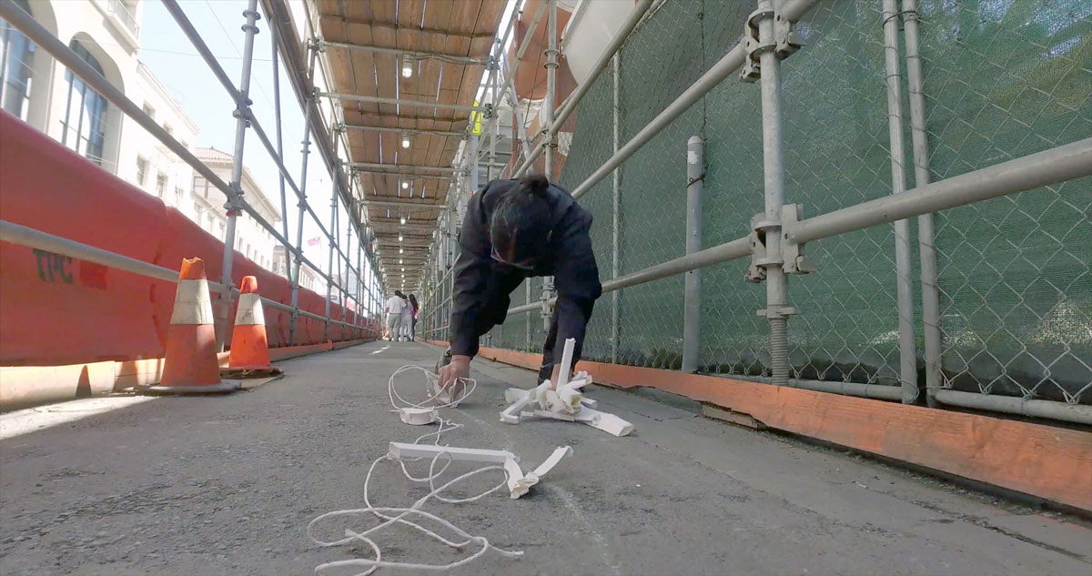 Figure bent over with white paper sculptures in hands underneath construction scaffolding