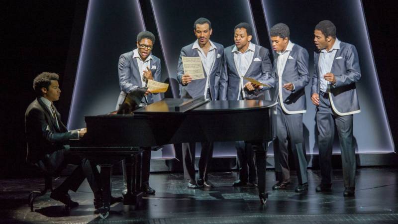 a group of men dressed as the Motown group the Temptations stand around a piano and sing