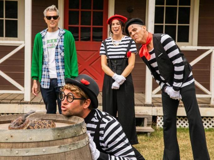 A man dressed as a mime goes face to face with a snake curled up on top of a barrel. Three figures watch on, two also dressed as mimes.