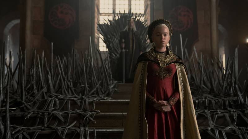 A woman in regal robes stands before a man on an iron throne.