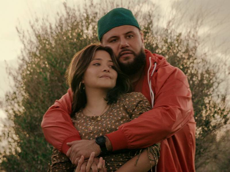 An Arabic man wearing a backwards baseball cap and red hoodie stands behind a Latina. He is embracing her warmly.