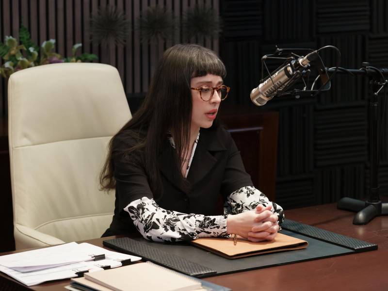 A woman sits at a desk, behind a microphone, hands clasped together on the desk before her. She is wearing spectacles and has long black hair with blunt bangs.