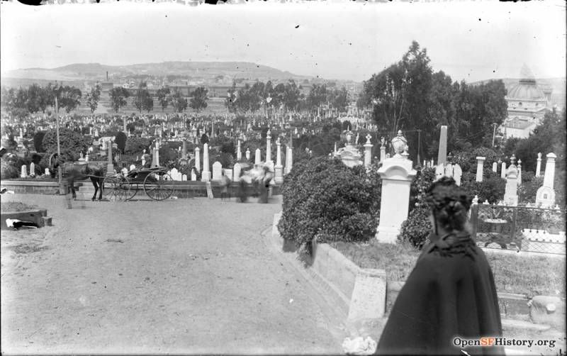 A woman in black Victorian mourning dress gazes down upon a graveyard full of marble monuments and gravestones.