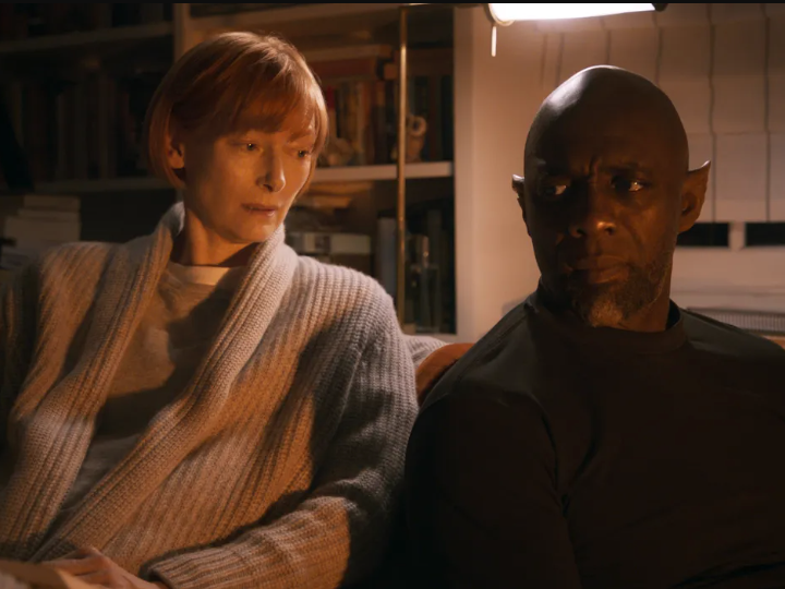 A white woman with short red hair sits on a couch next to a bald Black man with pointy ears.