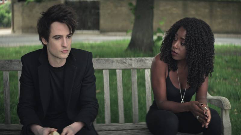 A young pale man sits pouting on a bench in a park. Next to him sits an attractive young Black woman, turning to see his face. Both are wearing all black clothing.