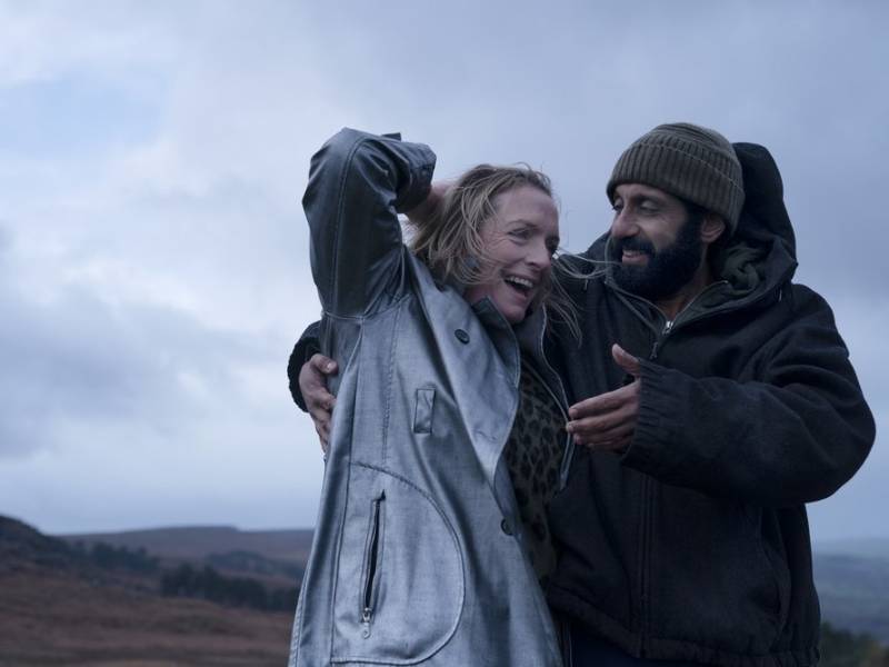 A middle-aged white woman laughs and reaches for the back of her head next to an smiling Pakistani man who is wearing a warm coat and beanie. They are standing on a hillside with grey skies behind them.