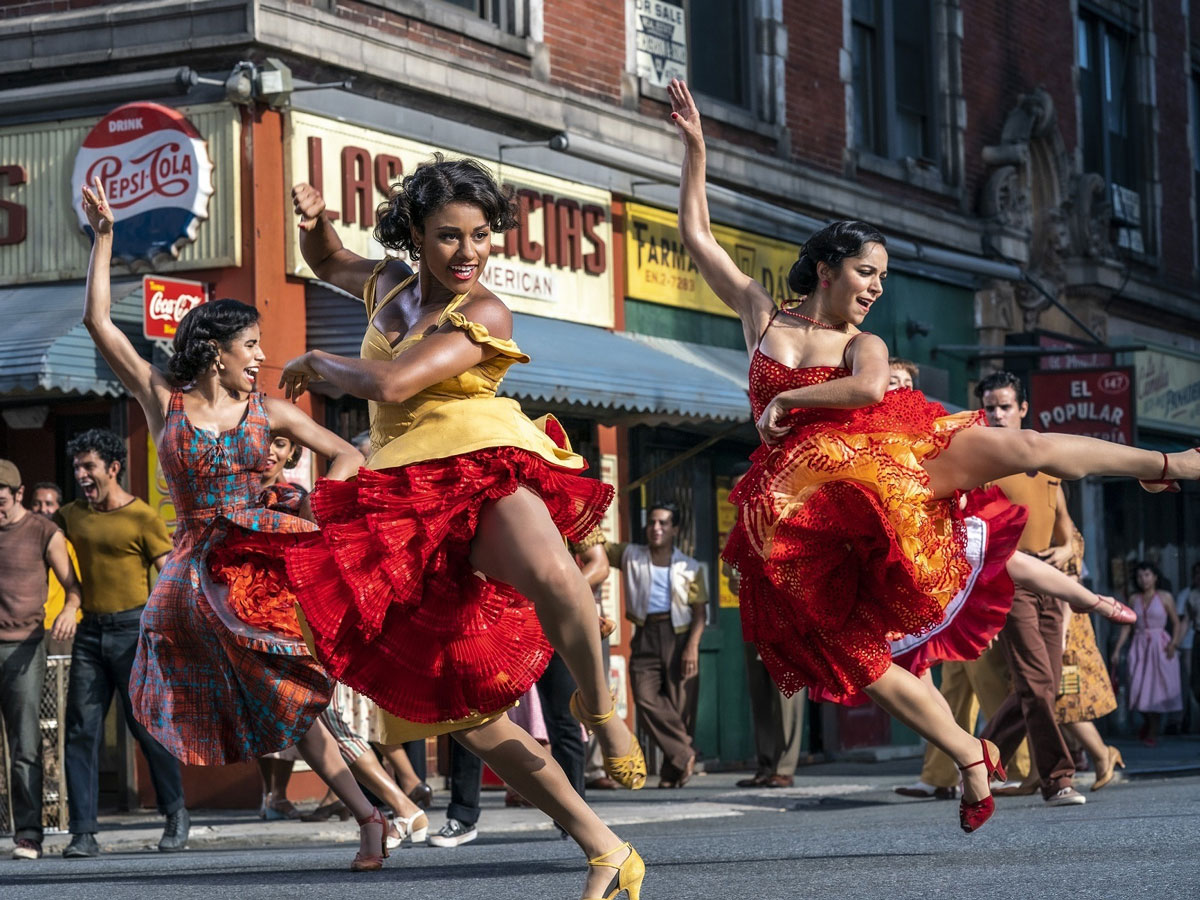 Three women in red, yellow and orange dresses dance expressively on the street outside a bodega.