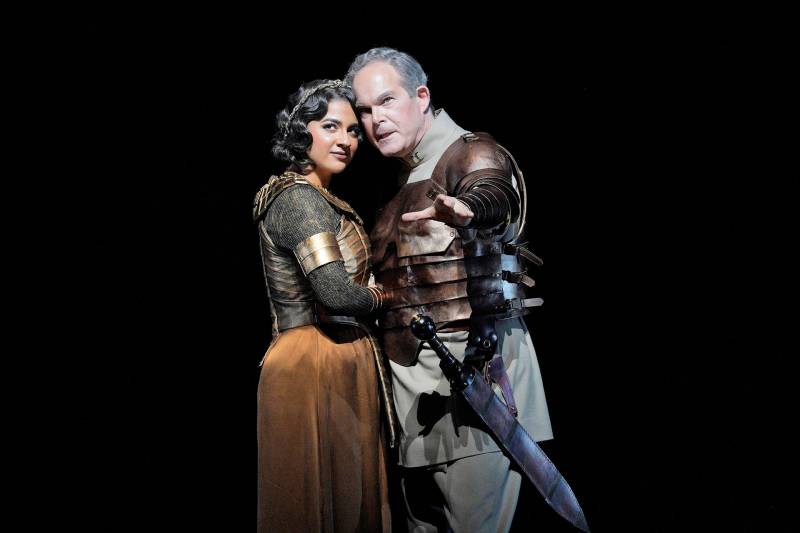 A woman and a man, both in armor, huddle close to each other, looking out toward the camera