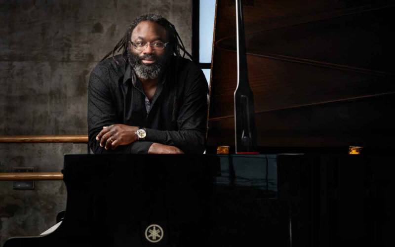 A Black man leans against a grand piano