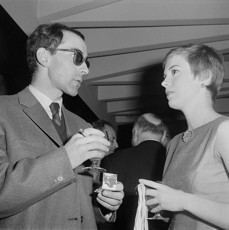 A white man in suit, tie and sunglasses stands with a woman in a dress.