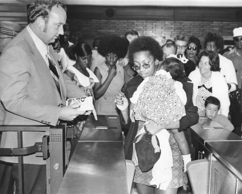 A Black woman carrying a toddler makes her way through BART barriers, ticket in hand, as crowds watch on.