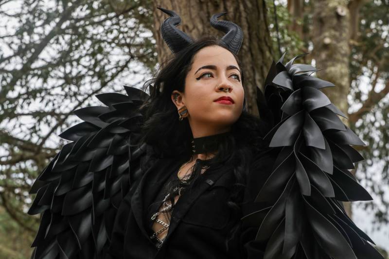A woman wearing black goat-like horns on her head and elaborate black wings stands firmly before a tree.