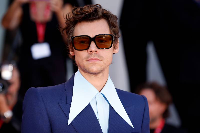Harry Styles, wearing a dark blue suit, stands on the red carpet, wearing large square sunglasses. He is also wearing a light blue shirt with aggressively long pointed collars.