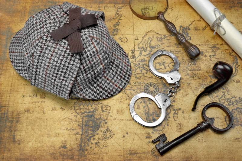 A stalking cap, handcuffs, magnifying glass, pipe, key and piece of rolled up paper lie on top of an old sepia-toned map.