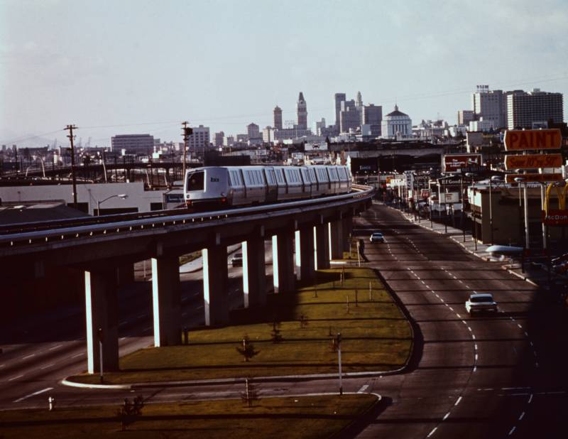 A train travels on elevated tracks, with the sparse Oakland skyline in the distance.