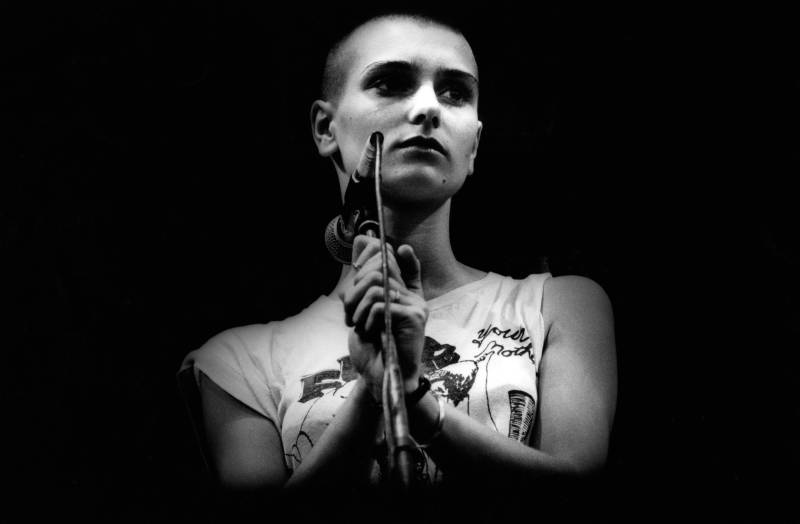 A woman with a shaved head stands behind a microphone holding the stand pensively as she looks off into the distance.