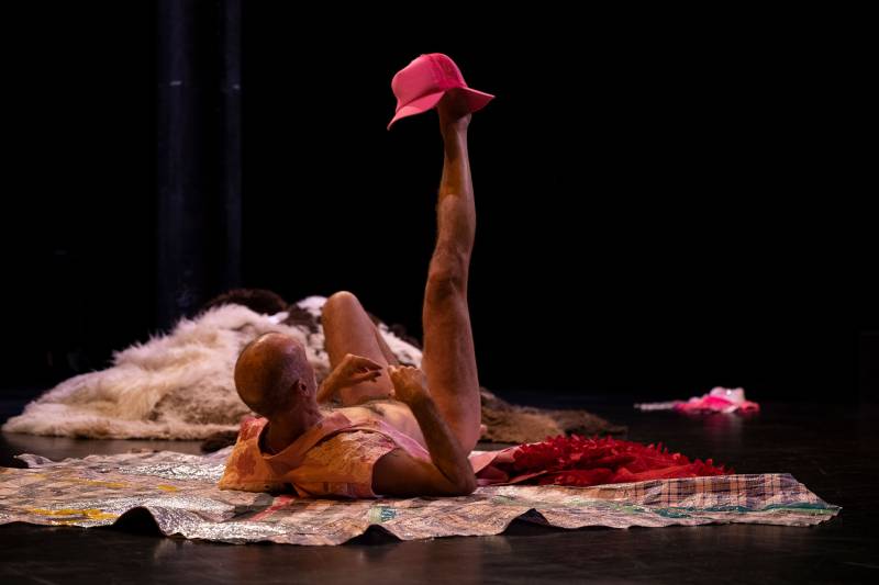 Keith Hennessy, an older white man, lays on his back in a theater nearly naked, wearing only a long pink vest, his right leg is lifted pointing to the sky and holding a hot pink hat with two bills. He lays on a sewn together quilt of reusable plastic shopping bags with a pile of sheepskins in the corner.