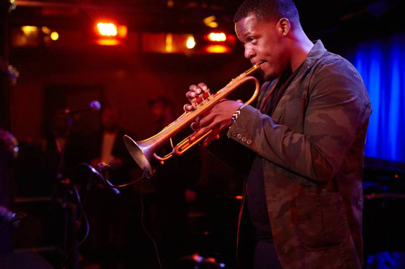 a Black man in a suit jacket plays the trumpet in a dimly lit club