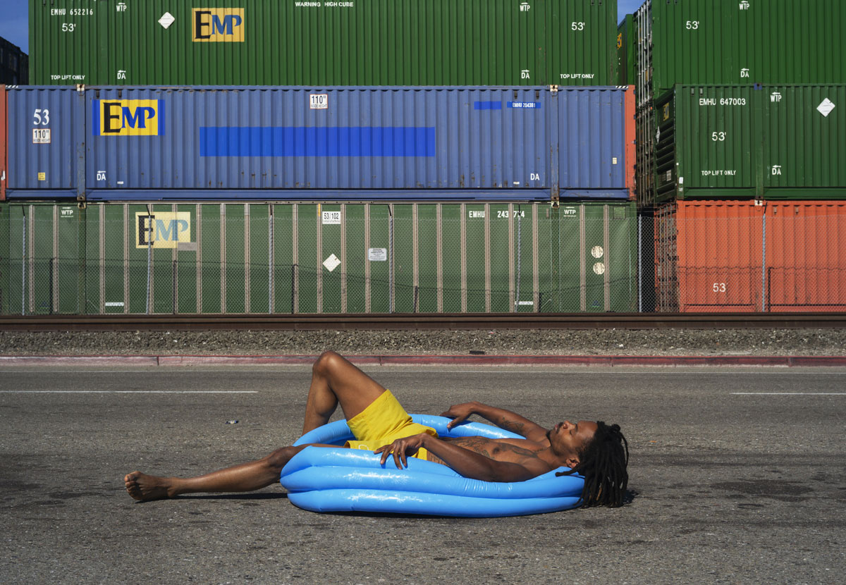Black man in swim trunks lays in kiddie pool in front of shipping containers