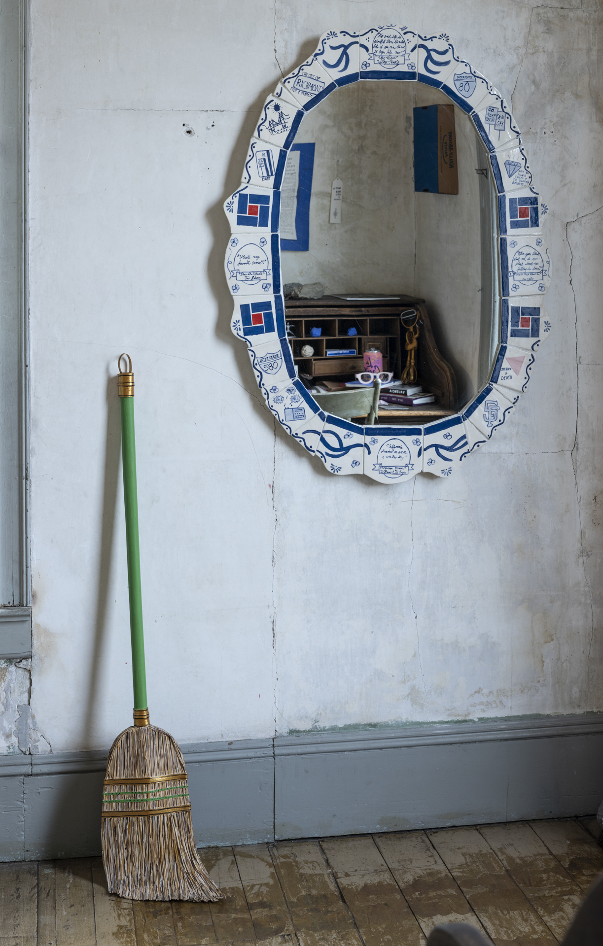 Paper painted broom leans against wall beside large oval mirror with cluttered desk reflected