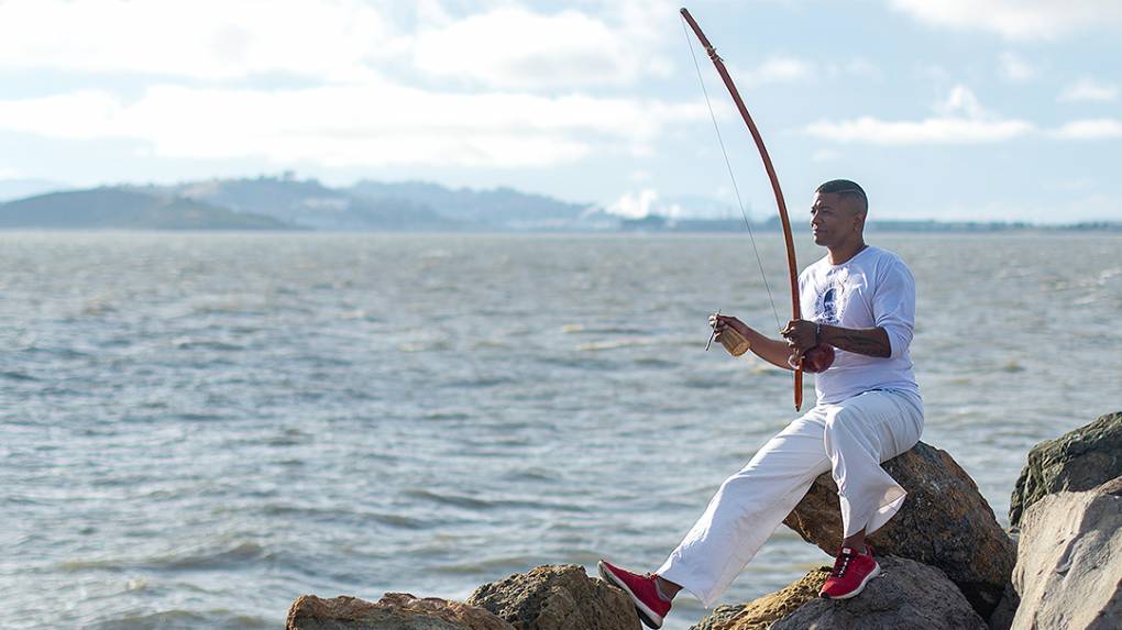 A capoeirista is sitting on a rock in front of the San Francisco Bay and is playing the berimbau.