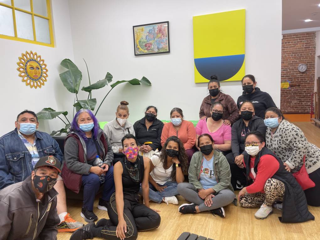 A group of 14 individuals are sitting and posing while wearing masks in what appears to be an art studio.