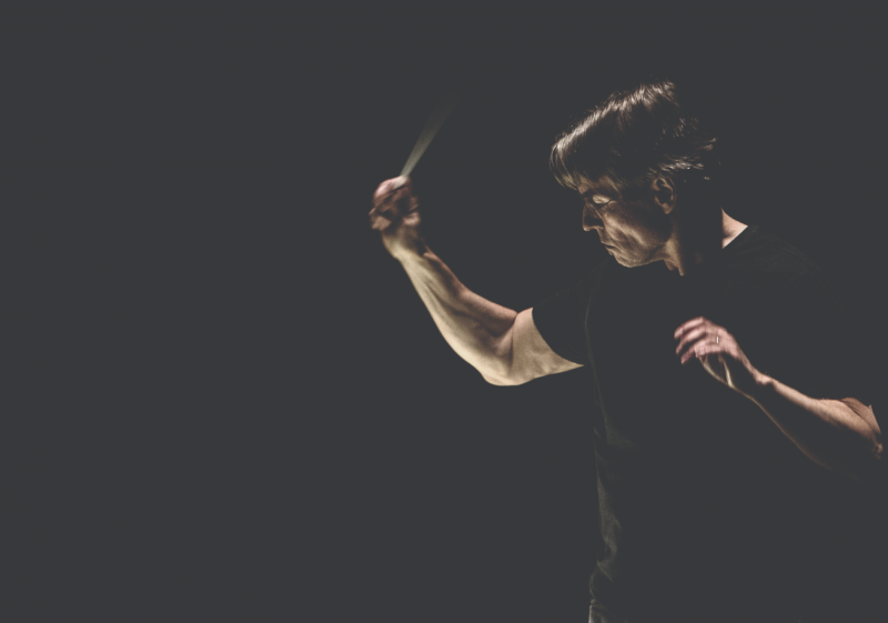A conductor in action, arm flexed out before him, before a black background. He is wearing a casual black t-shirt, rather than a suit.