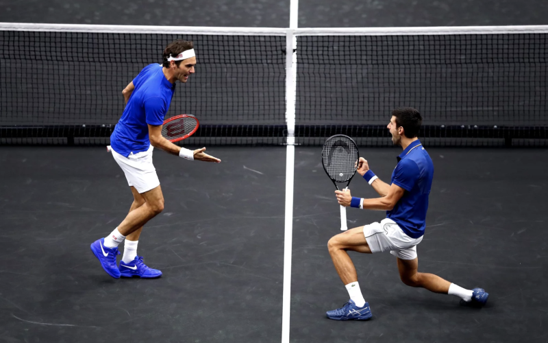 Two tennis players on the same side of the net wearing matching blue t-shirts and white shorts. They are gesturing in celebration.