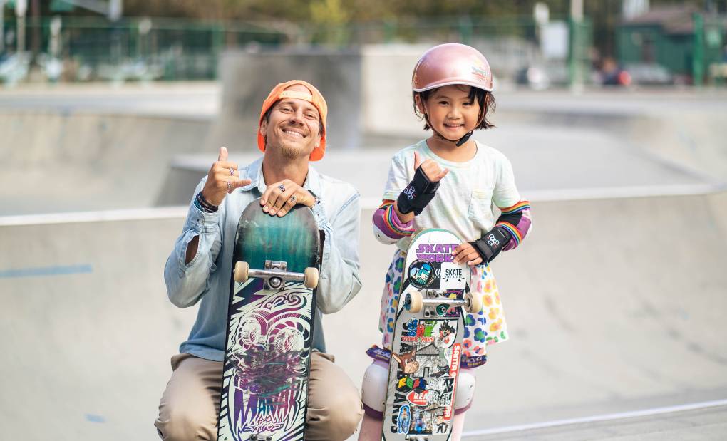 a man and a 6-year-old girl both pose holding skateboards at a skate park