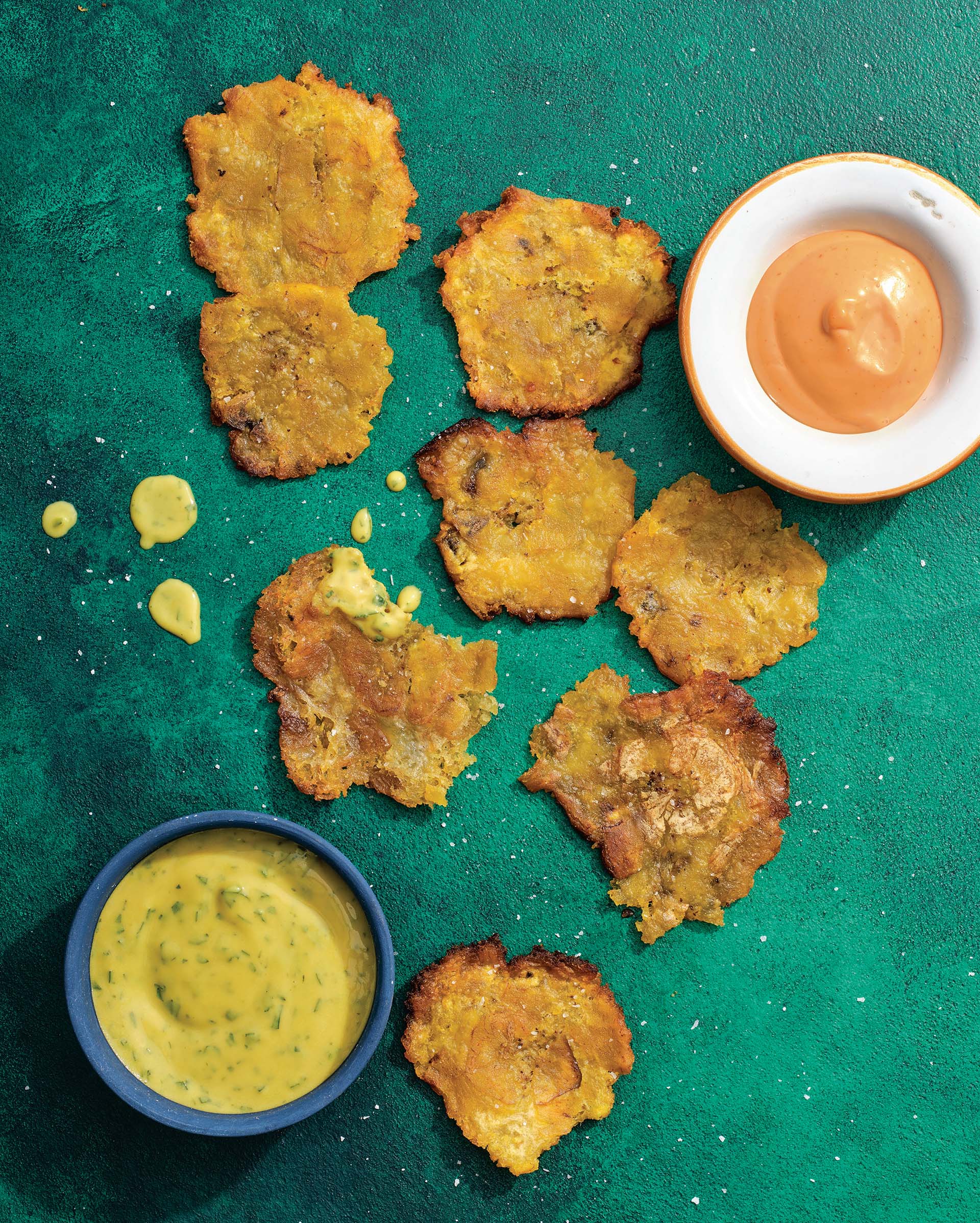 Overhead shot of thin, crispy tostones against a green backdrop, with two dipping sauces on the side.