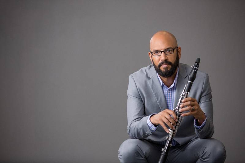 A Black man with bald head and neat beard wearing a grey suit and holding an oboe.