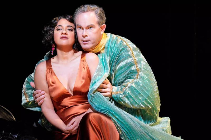 a woman in an orange dress and a man in a green robe embrace against a black backdrop on stage