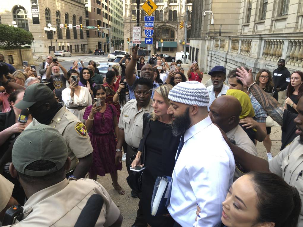 A crowd of people gathered in the street, stands in a circle around a bearded man of Arabic descent and a white woman.