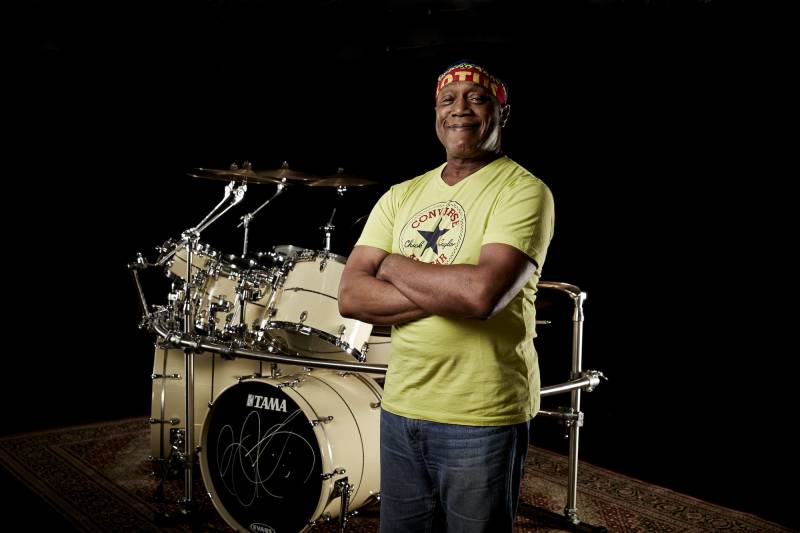 a Black man in a Converse All-Stars t-shirt stands on front of a drum set