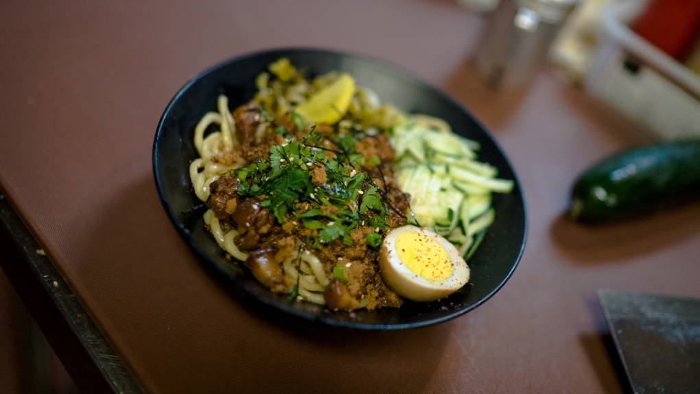 A bowl of noodles topped with meat sauce, slivered cucumbers and half a hard-boiled egg.
