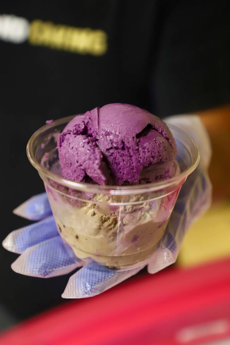 the owner of Ellis Creamery holds up a clear cup of purple ube ice cream with an extra scoop of cookies-and-cream