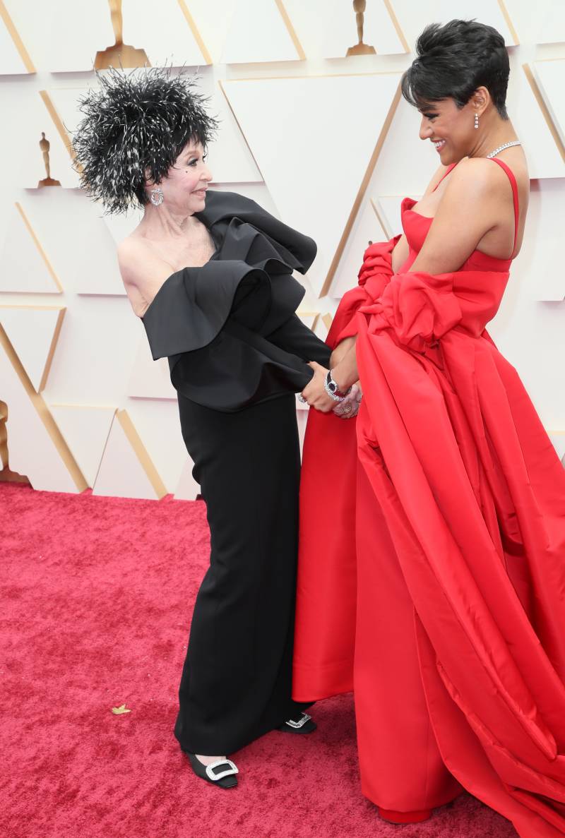 Two women, wearing elaborate evening gowns stand face to face and gaze warmly at each other on the red carpet.