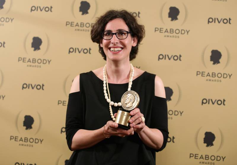 A woman wearing black dress, white pearls and black spectacles smiles as she holds up a trophy.