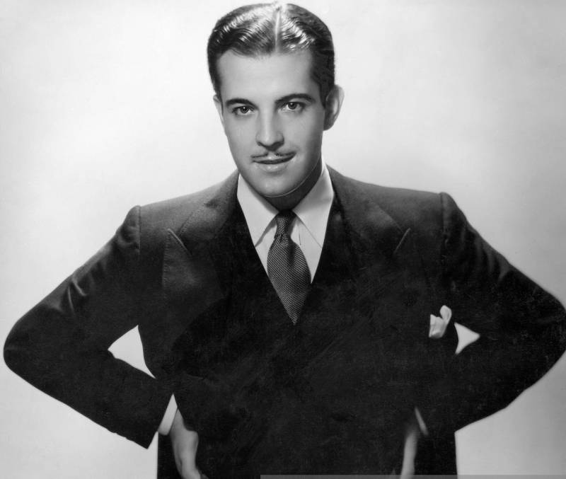 A suave man with slicked back hair and a pencil mustache stands with his hands on his waist, wearing a sharp suit and tie.