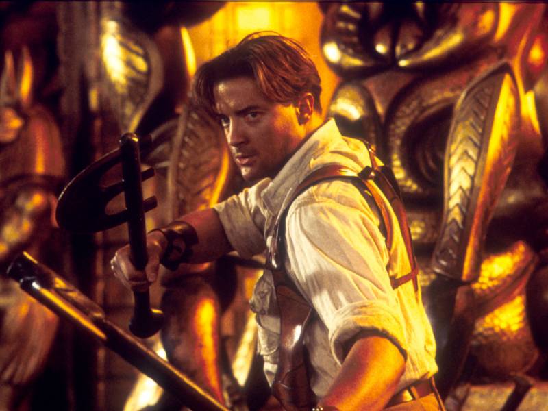 A dashing young man wearing a white shirt, sleeves rolled up, stands poised for action, two ancient-looking weapons held out in front of him.