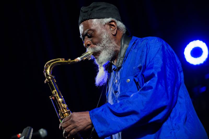 An elderly African American man in a blue shirt and long white beard plays the tenor saxophone