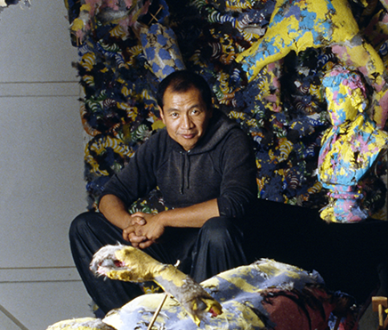 A Filipino American man dressed in black pants and hoodie sits low to the ground on the edge of a surreal sculpture. 
