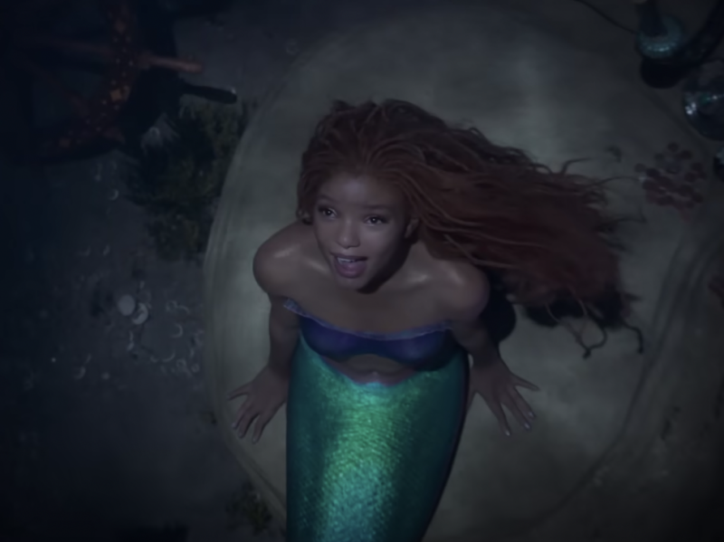A mermaid sings underwater while sitting on a rock. She is a beautiful young Black woman with flowing red hair and a green tail.