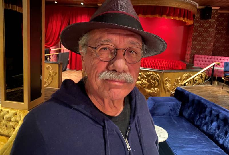 An older man with a grey mustache and spectacles stands in a grand ballroom, wearing a hoodie and a fedora.