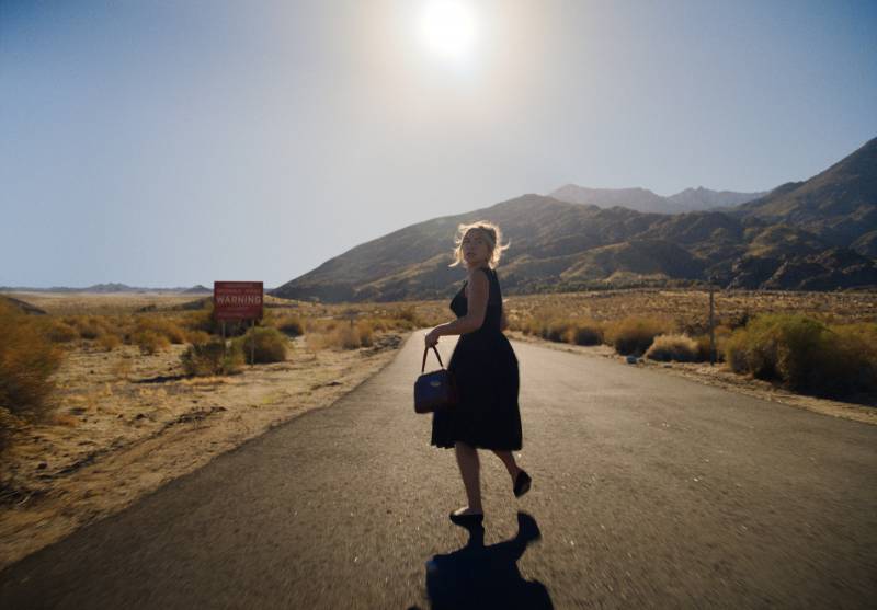 A petite blond woman wearing a pretty black 1950s-style dress runs in the center of a road towards desolate mountains. She is glancing back over her shoulder.