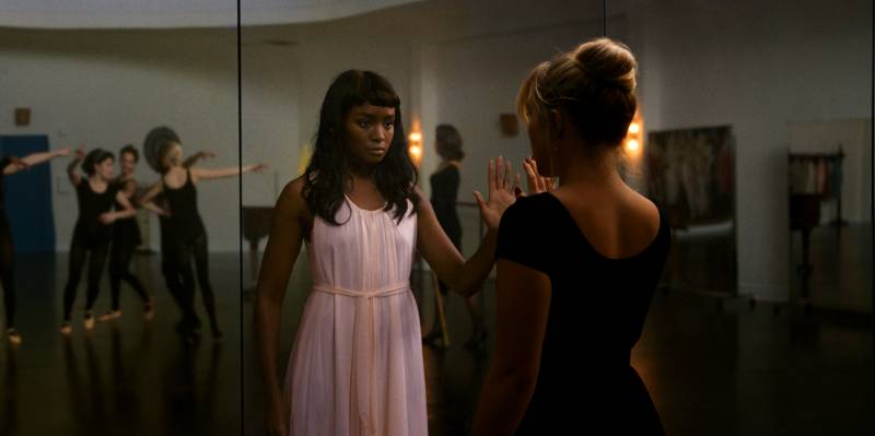 A white woman in a black leotard stares at her reflection in the mirror. Staring back at her is a pretty Black woman with long straight hair, wearing a pink nightgown.