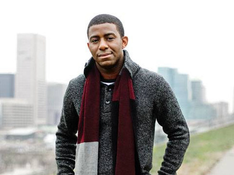 A young Black man stands outdoors on a Fall day. A city skyline is visible behind him.