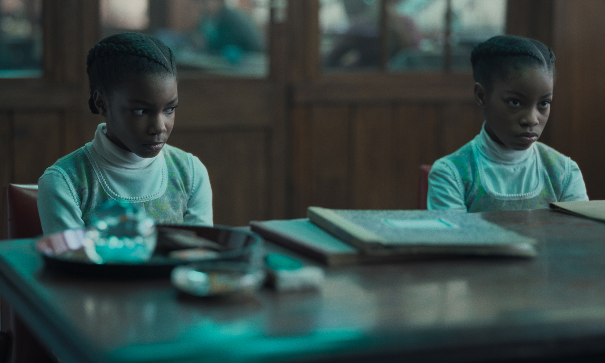 Two young Black girls in matching outfits sit behind a large table; one glances at other