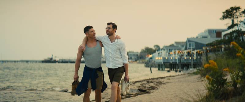 Two men walk along a beach with their arms around each other.