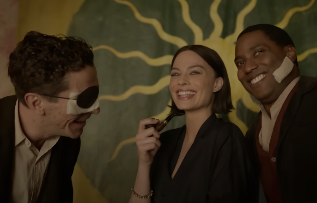 A man with an eye patch leans forward next to a white woman smoking a pipe and a Black man with a bandage on his face. All three of them are laughing.
